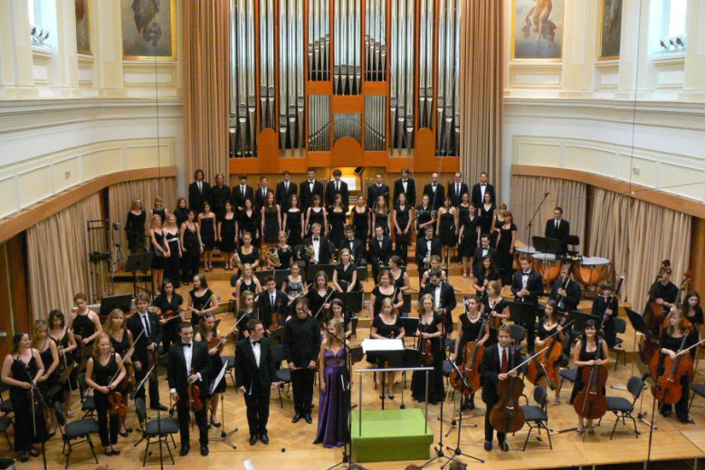 Moški in ženske z instrumenti in pevci ter dirigent stojijo na odru v Slovenski Filharmoniji