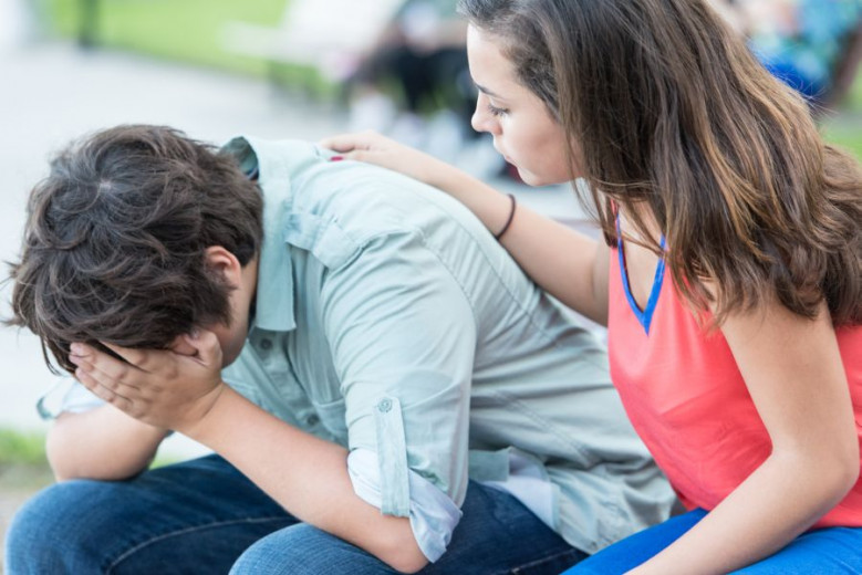 The teenage boy is crying with his hands in his face and the teenage girl is comforting him.