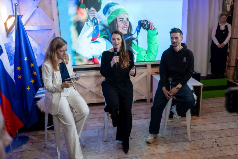 Three people are seated on stools in front of a screen displaying a jubilant athlete holding a medal. A woman in white holds a tablet, a woman in black holds a microphone, and a man in a hoodie sits casually. Slovenian and EU flags are in the background, suggesting a formal event. The atmosphere is celebratory and engaged.