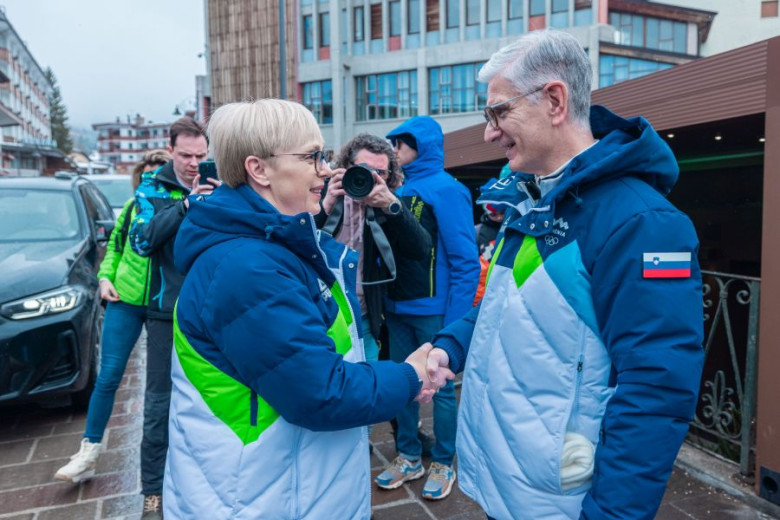 Two people in winter jackets with Slovenia flags shake hands outdoors, smiling. Photographers capture the moment, creating a cordial and lively atmosphere.