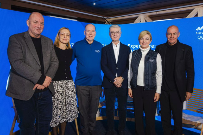 A group of six individuals stands in front of a blue background with Olympic logos. They are dressed in business or casual attire, smiling and posing for the photo.