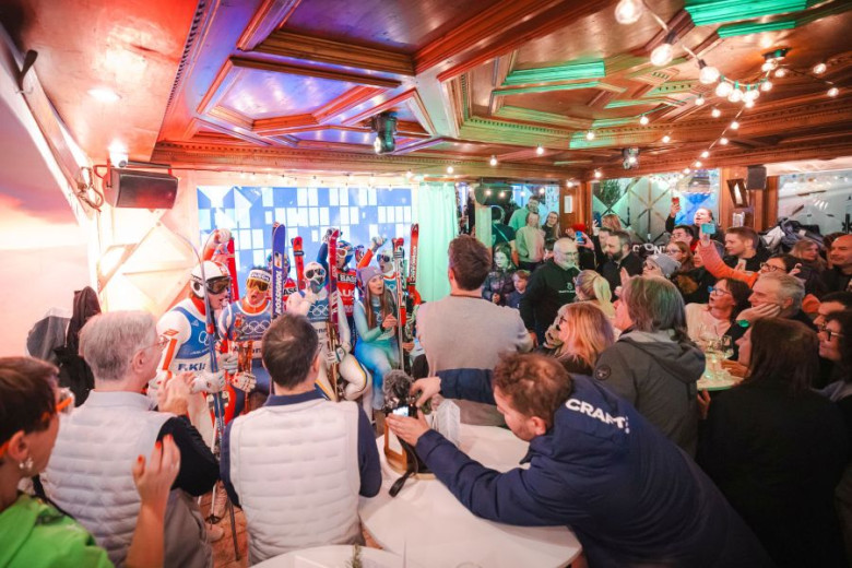 A lively crowd in a rustic bar gathers around a group of people in colorful skiing gear, holding skis. The mood is festive and energetic.