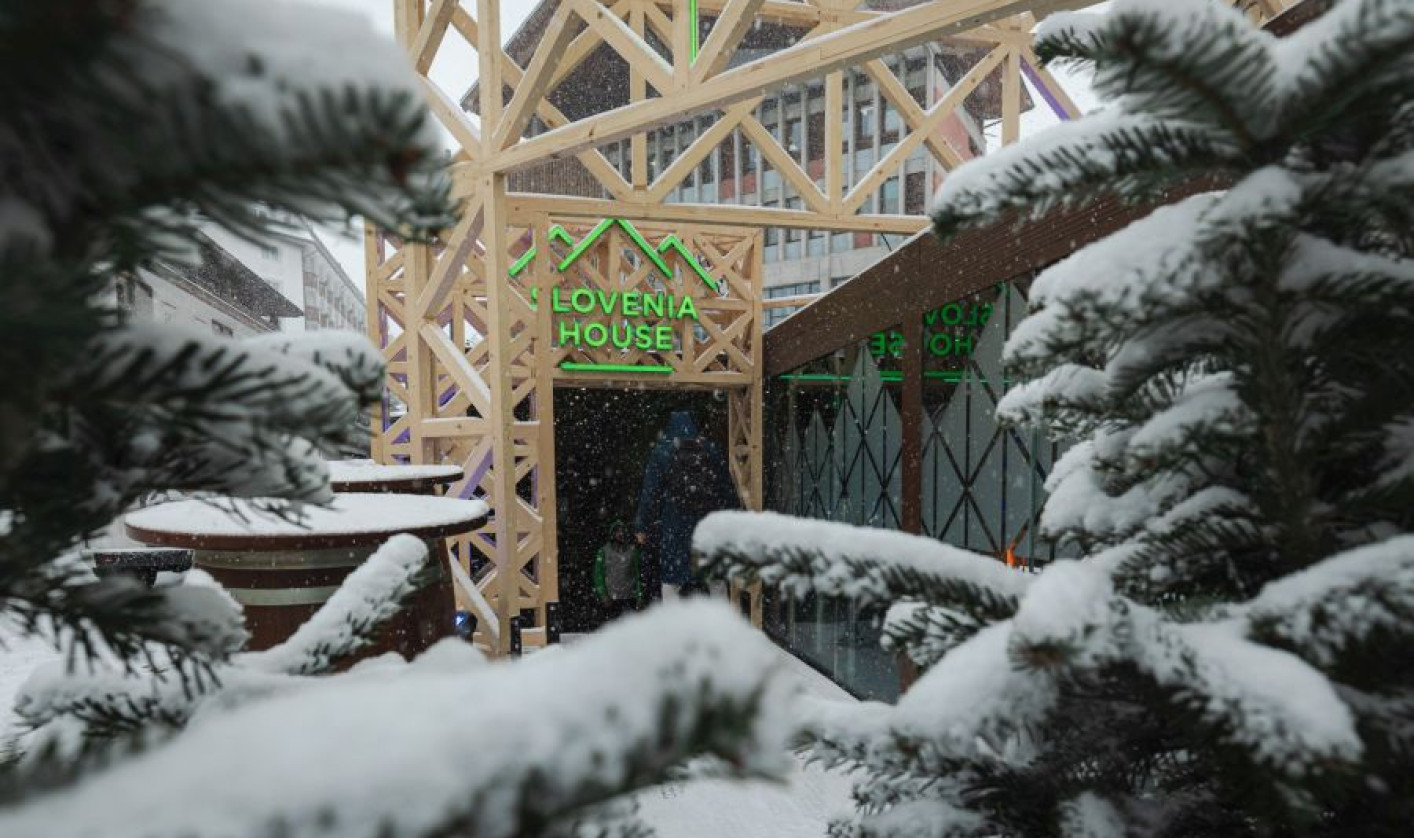 Snow falls gently on evergreen trees framing a wooden structure labeled "Slovenia House." The scene is quiet, conveying a wintery, peaceful ambiance.