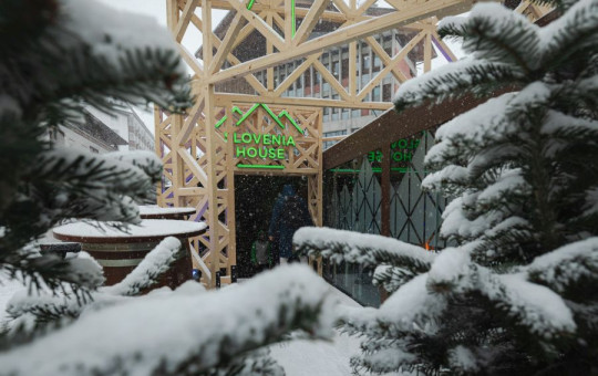Snow falls gently on evergreen trees framing a wooden structure labeled "Slovenia House." The scene is quiet, conveying a wintery, peaceful ambiance.