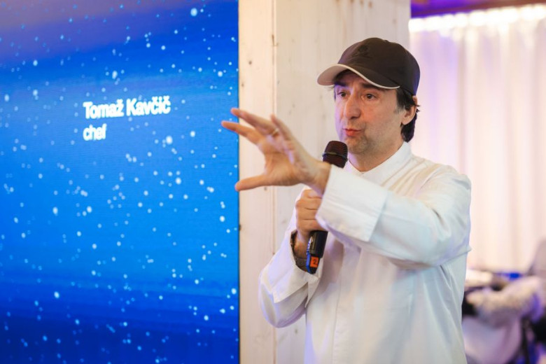 A chef in a white coat and cap speaks passionately into a microphone, gesturing with his hand. A screen displaying his name is visible in the background.