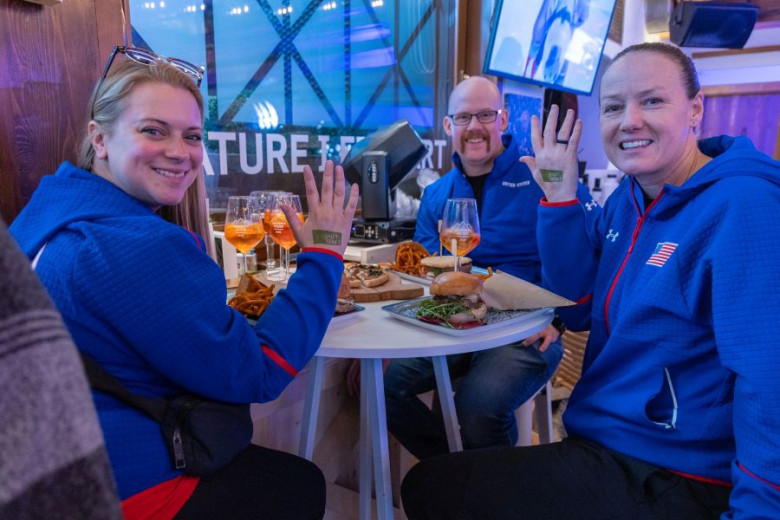 Three people in blue jackets smile at a cafe table with drinks and food, happily showing hand stamps. The atmosphere is lively and relaxed.