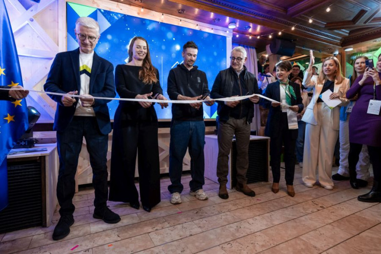 A group of diverse individuals smiles while holding a ribbon, about to cut it at an indoor event. A European flag is partially visible on the left.