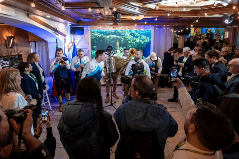 A lively band plays traditional instruments in a cozy, warmly-lit room filled with an engaged crowd. People capture the moment with cameras and phones.