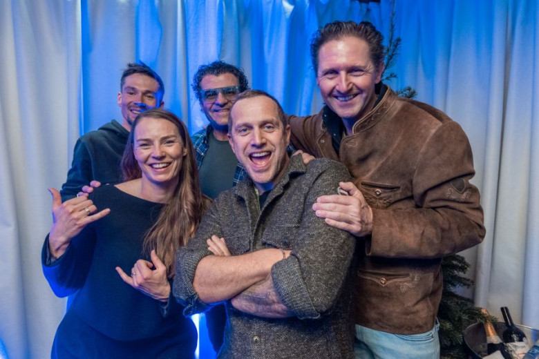 A group of five people smiling and posing together in front of blue-lit curtains. They appear cheerful and are wearing casual, warm clothing.