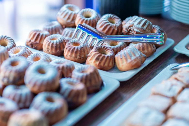 A platter of small, powdered bundt cakes with a shiny metal tong resting on top, creating a tempting display of pastries. Bright, inviting atmosphere.