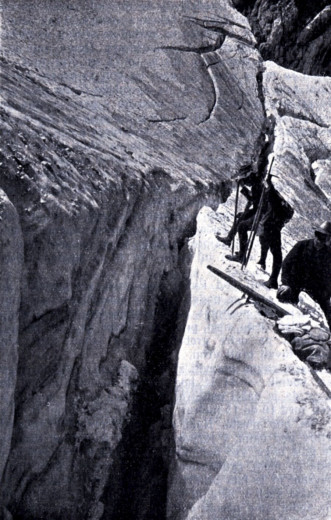 Old, black and white, photo shows mountaineers walking at the edge of a deep Skuta Glacier crack. 