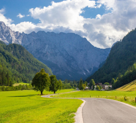 A winding country road curves through vivid green alpine meadows toward a village, framed by forested slopes and gray mountains.