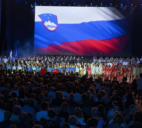 A large group of adults and children performs on an outdoor stage in front of a large audience. The backdrop displays the Slovenian flag.