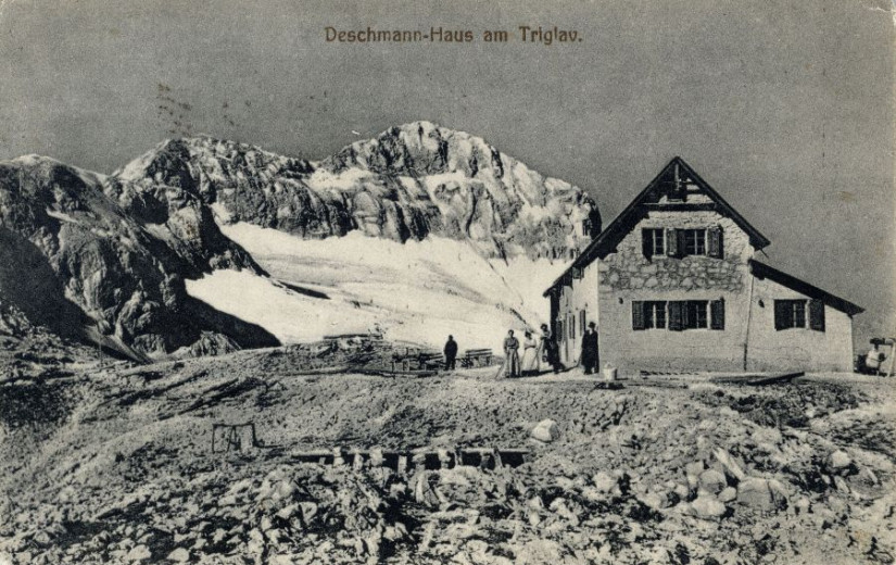 Old, black and white, postcard from 1910 showing a mountain lodge with people standing next to it and a steep, rocky mountain slope, mostly covered with snow in the backgroud.nd 