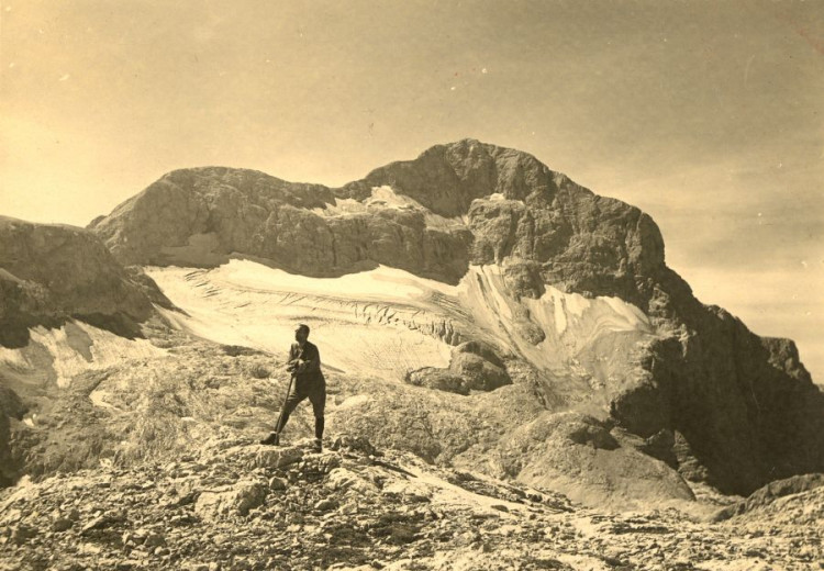 An old photo shows a man standing in the mountains with steep mountain slopes in the background. The lower parts of the slopes in the background are covered with snow and ice.