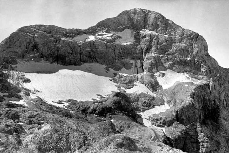 A black and white photograph of a steep mountain slope scattered with snow patches of varying sizes.