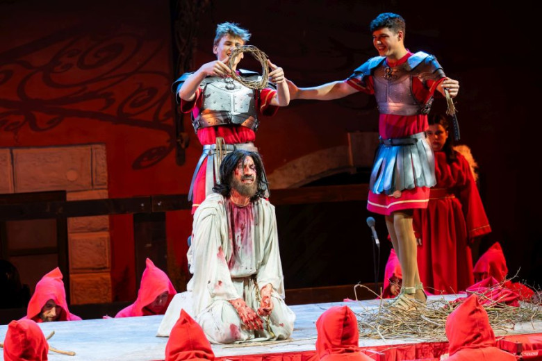 Three actors are recreating the Crowning with Thorns scene on a small wooden platform which is being carried outdoors in the street by men in red cloaks with pointed hoods.