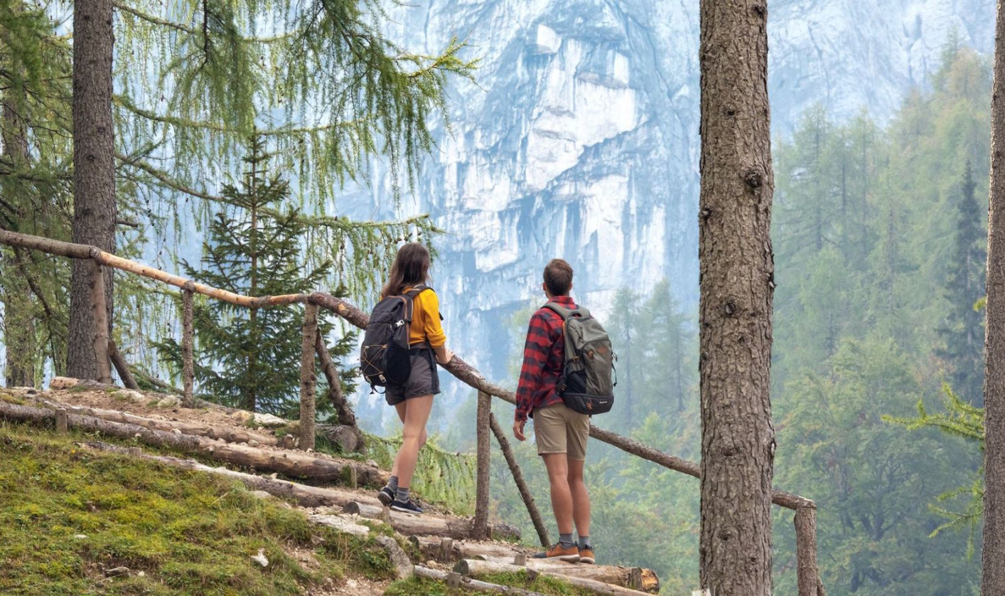 Two hikers with backpacks pause on a tree-lined path, gazing at distant rocky cliffs. The scene conveys exploration and appreciation of nature.