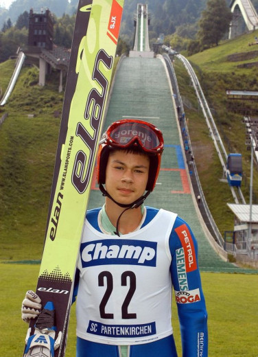 Young ski jumper in a helmet holds skis bellow of a ski jump, with a green ramp and hills behind.