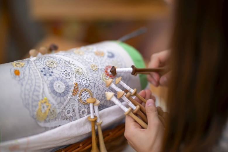 A close-up of a woman, holding wooden sticks "klekeljni", and crafting colorful bobbin lace on a bobbin lace pillow.