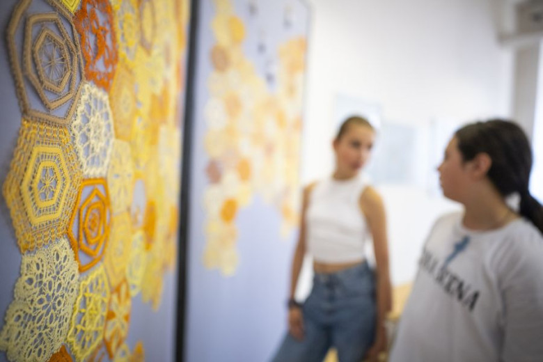 Blurred photograph of a yellow, orange and brown lace beehive, hanged on a wall, and two girls talking in front of it. 