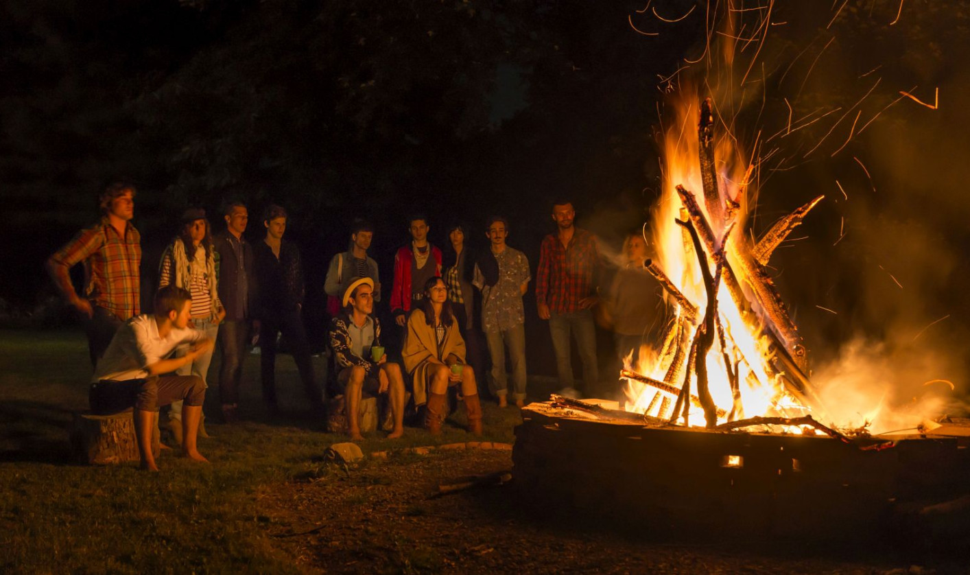 A burning bonfire and a group of people sitting and standing in halfcircle in the background.