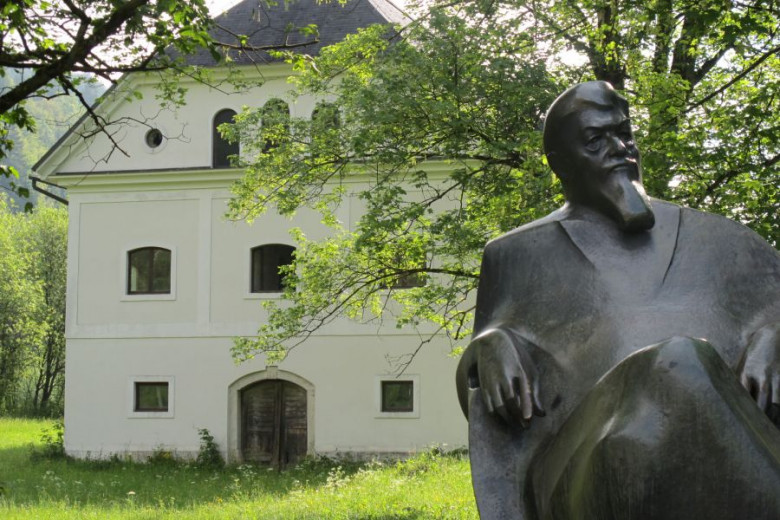 Hiša, travnik, v ospredju stoji kip Ivana Tavčarja.