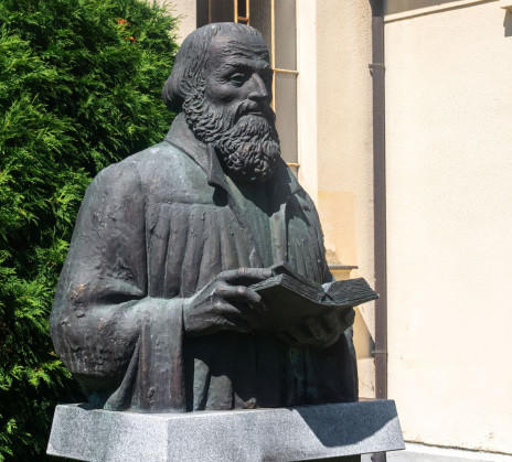A bronze bust statue of Primož Trubar, holding an open book, in front of a building.