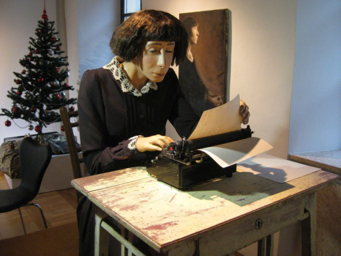 A mannequin with a short bob haircut and vintage clothing types on a typewriter at a rustic desk. A decorated Christmas tree and portrait are in the background.