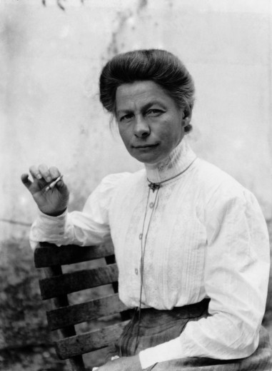A woman in early 20th-century attire sits on a bench, wearing a high-collared white blouse. She holds a cigarette, exuding a calm, confident demeanor.