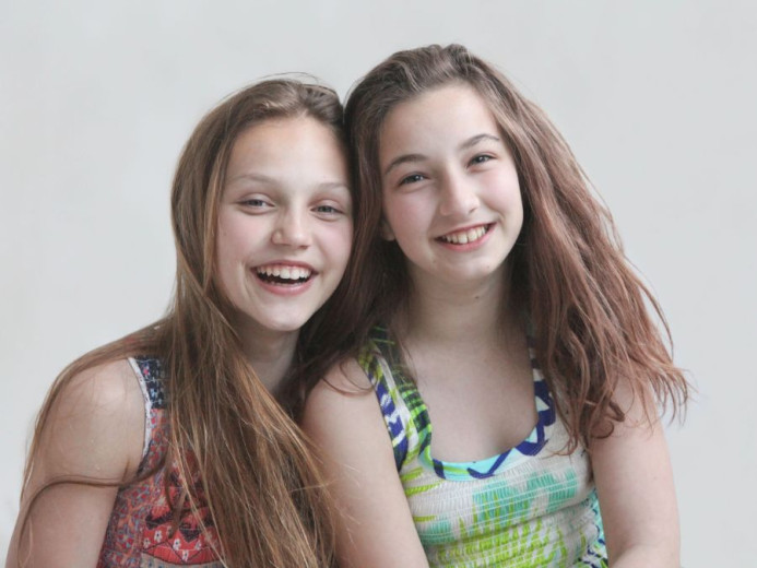 Two young girls with long hair smile brightly at the camera. One wears a colorful patterned top, the other a green and blue tank. They convey joy and friendship.