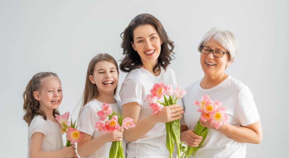 A joyful group of four women across different generations, wearing white shirts, hold pink flowers against a light gray background, conveying warmth and unity.