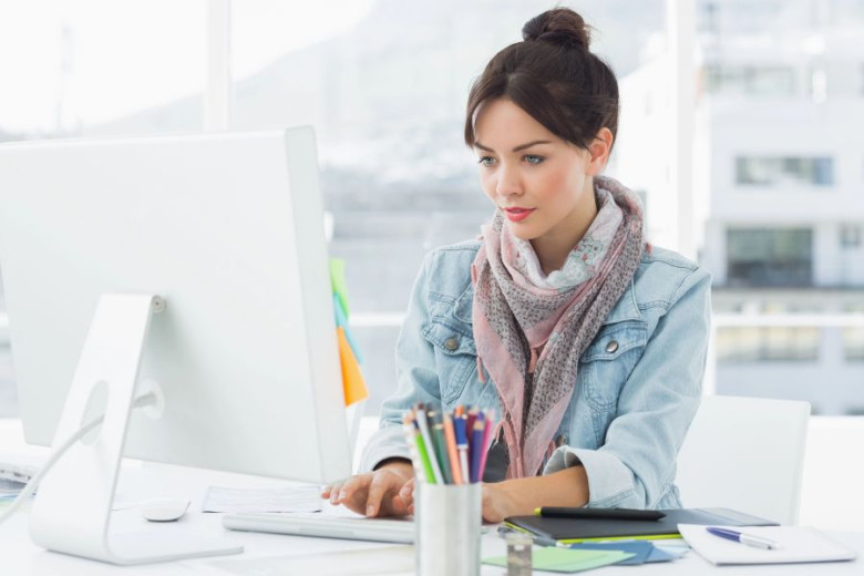 Young woman with dark hair in a bun and scarf focuses on a computer in a bright office. She appears attentive, with notepads and colorful pens nearby.
