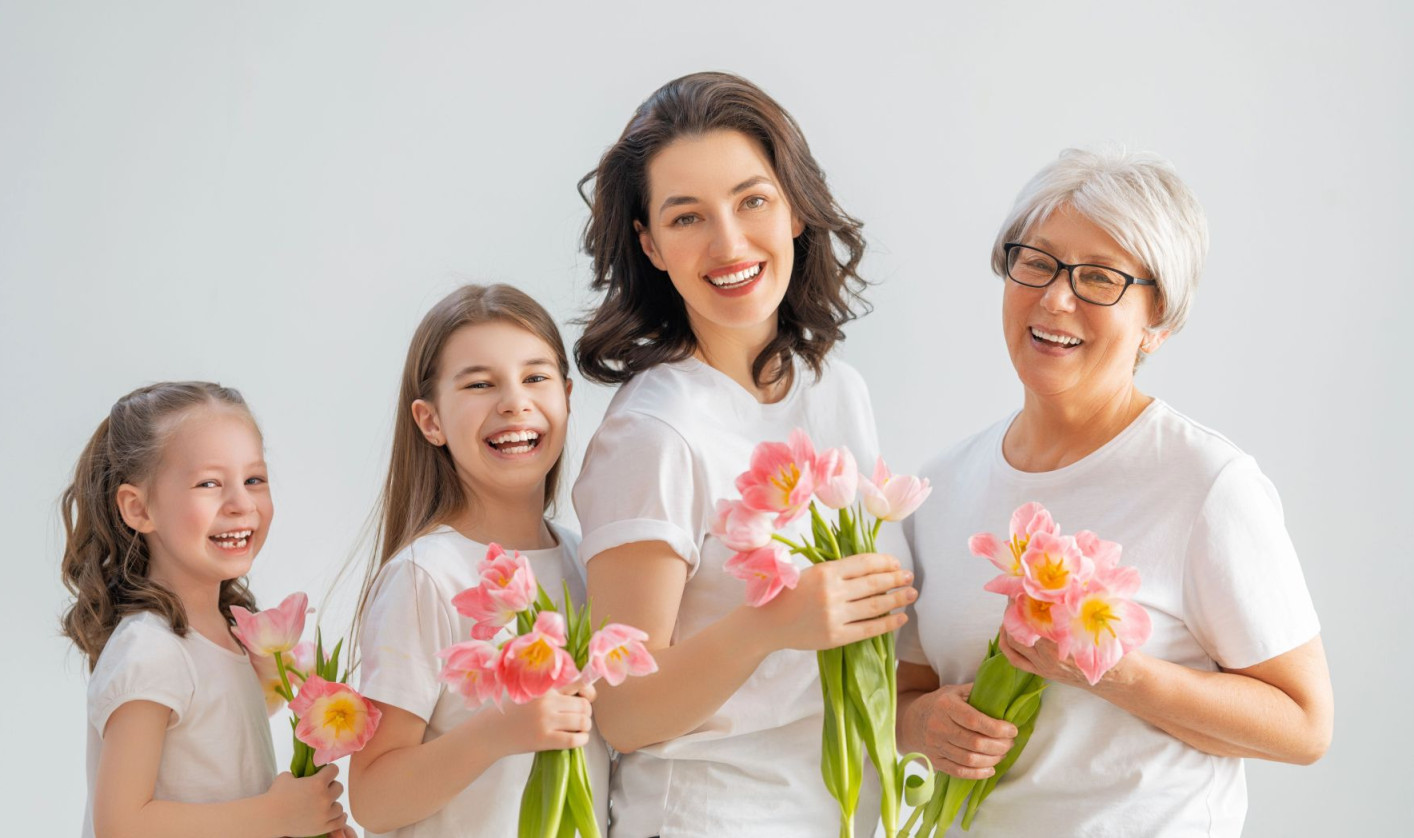 A joyful group of four women across different generations, wearing white shirts, hold pink flowers against a light gray background, conveying warmth and unity.