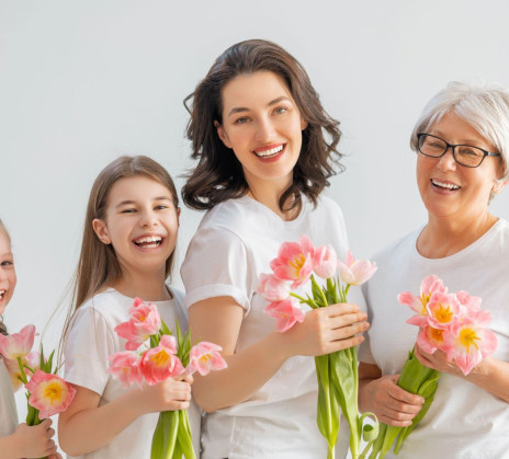 A joyful group of four women across different generations, wearing white shirts, hold pink flowers against a light gray background, conveying warmth and unity.