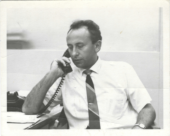 Anton Banko in a white shirt and tie sits at a desk and talks on the phone.