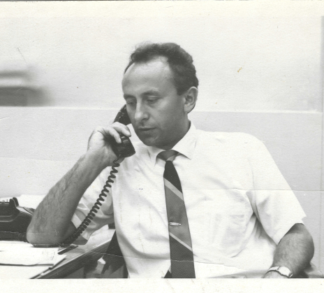 Anton Banko in a white shirt and tie sits at a desk and talks on the phone.