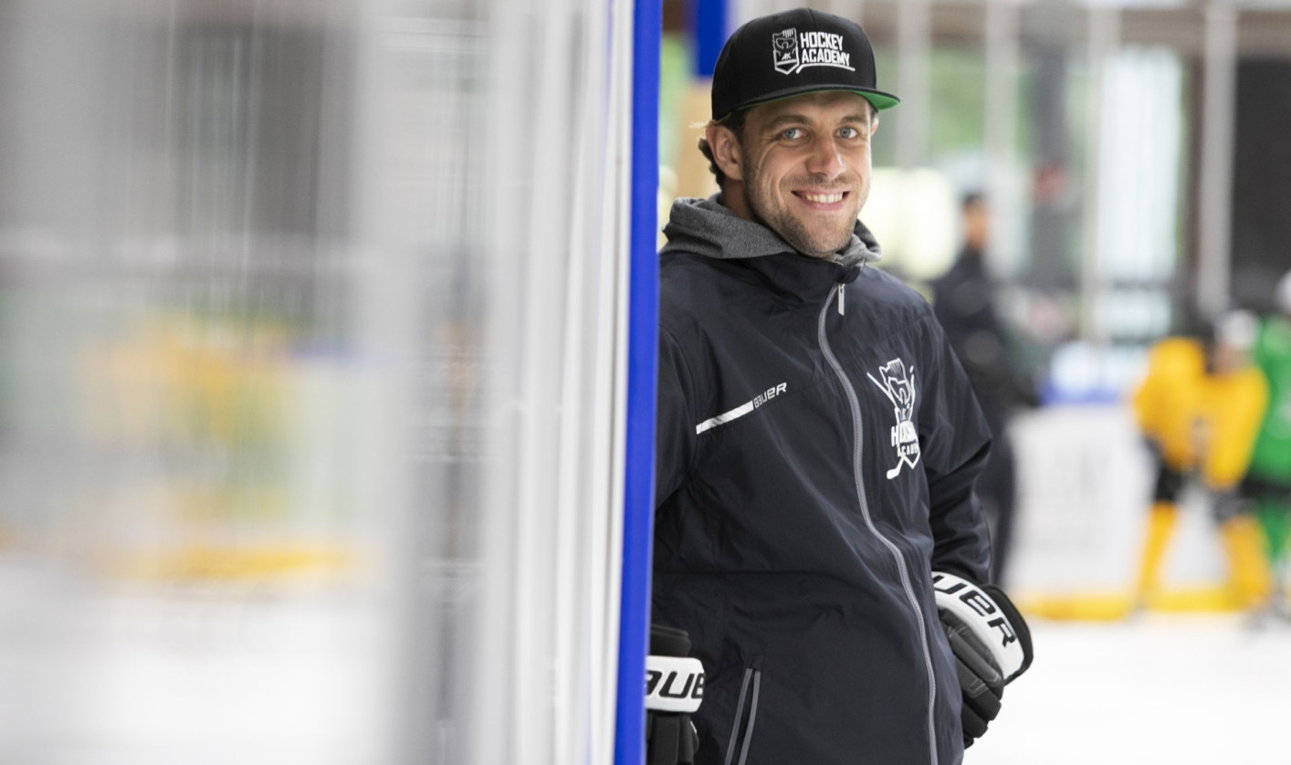 A portrait photo of Anže Kopitar in black sports clothes and a cap, smiling and standing at the border of the ice rink.