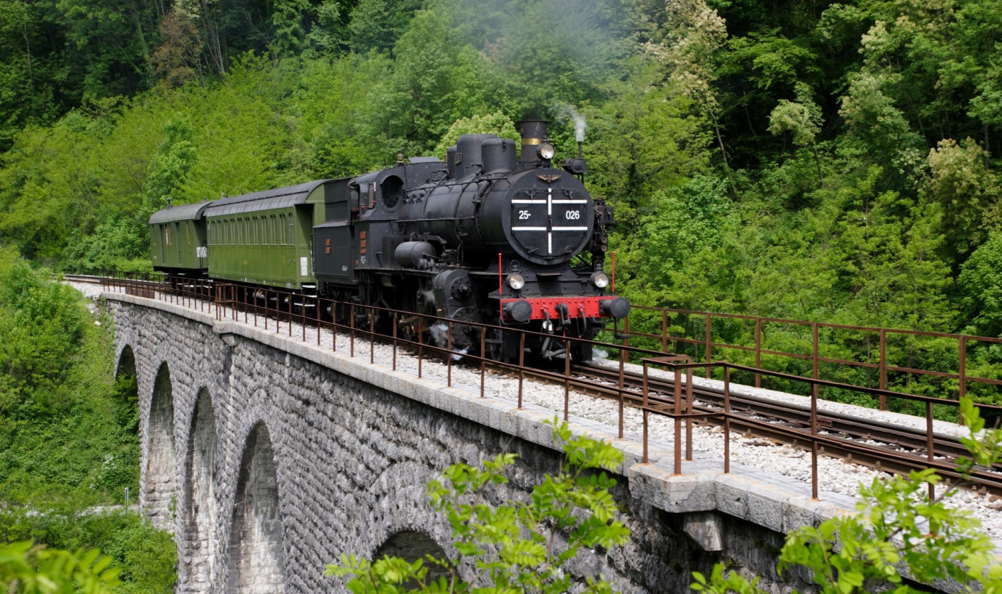 An older black steam locomotive is driving on a railway across a stone arch bridge, surrounded by a rich green forest. 