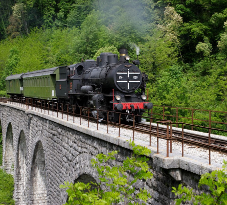 An older black steam locomotive is driving on a railway across a stone arch bridge, surrounded by a rich green forest. 