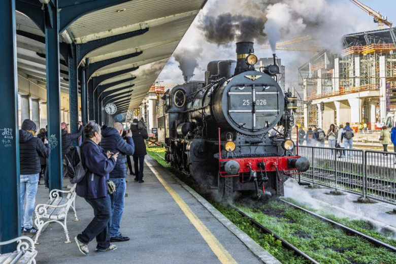 A black steam locomotive stands at a train platform. White steam rises from the engine. People are standing nearby, some taking photos. The platform has a roof, benches, and metal columns. Construction cranes and unfinished buildings are visible in the background.