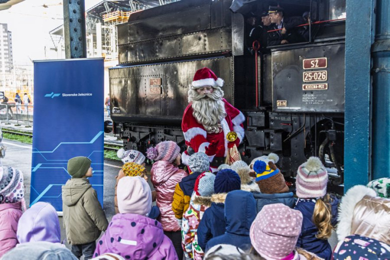 Santa Claus stands on a train platform in front of a black steam locomotive. He wears a red suit and holds a small bell. In front of him is a large group of young children dressed in winter jackets, hats, and backpacks. The children are facing Santa and listening to him. A train worker is visible inside the locomotive cabin. A blue sign with railway branding stands to the left.