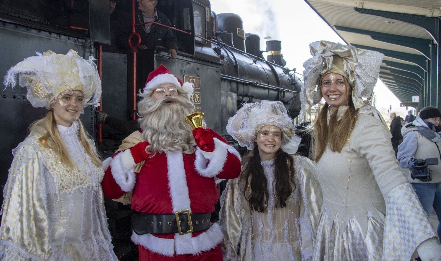 Four people stand on a train platform in front of a black steam locomotive. In the center is Santa Claus wearing a red suit, white beard, and black belt, holding a small golden bell. On both sides of him are three women dressed as angels in long, light-colored dresses with decorative hats. They are smiling and facing the camera. A train driver is visible inside the locomotive cabin behind them. The platform has a roof with metal columns, and other people stand in the background.