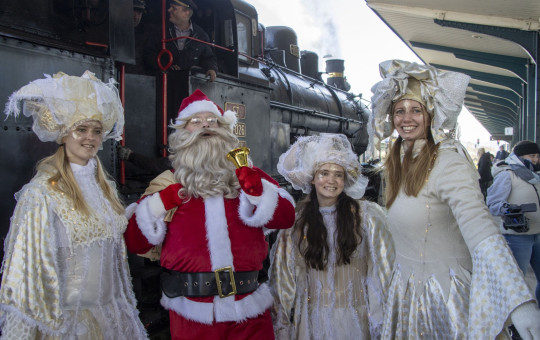 Four people stand on a train platform in front of a black steam locomotive. In the center is Santa Claus wearing a red suit, white beard, and black belt, holding a small golden bell. On both sides of him are three women dressed as angels in long, light-colored dresses with decorative hats. They are smiling and facing the camera. A train driver is visible inside the locomotive cabin behind them. The platform has a roof with metal columns, and other people stand in the background.