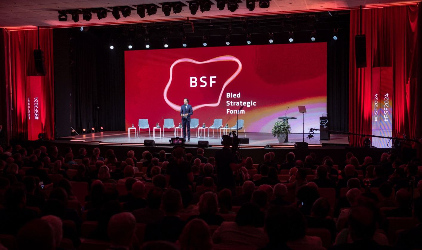 A stage at a BSF event with a speaker, 5 chairs lined up behind him and a projection of the BSF logo on a screen. A crowd of people sitting on chairs looking at the stage.