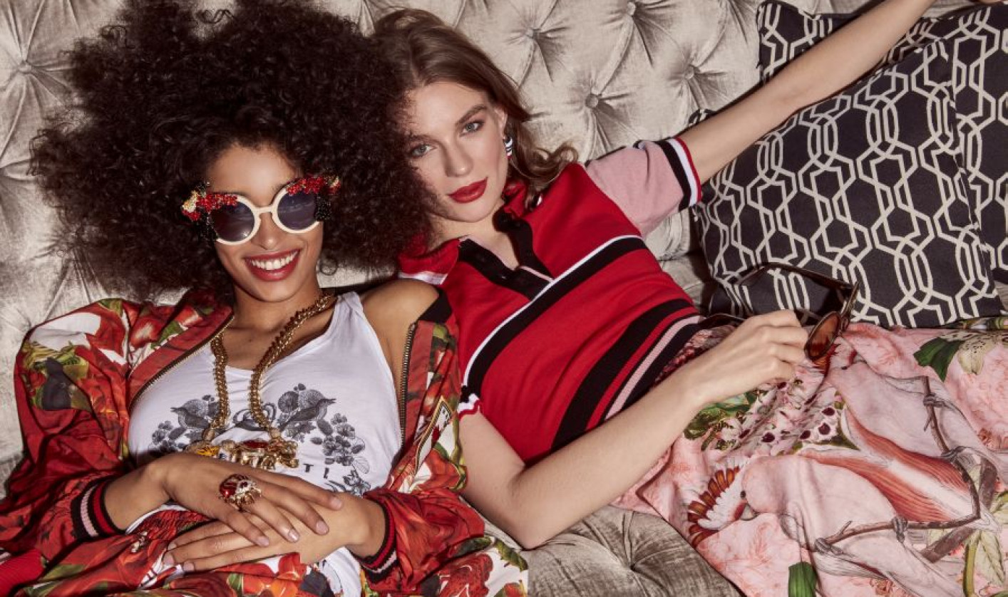 Two girls, where one is wearing sunglasses, are lying on a silver couch, wearing red, white and pink clothes, and smiling at the camera.