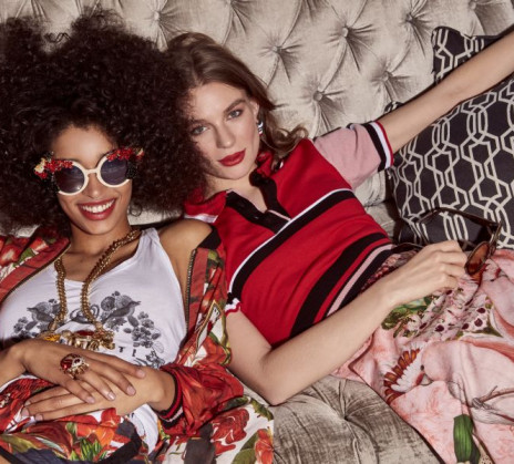 Two girls, where one is wearing sunglasses, are lying on a silver couch, wearing red, white and pink clothes, and smiling at the camera.