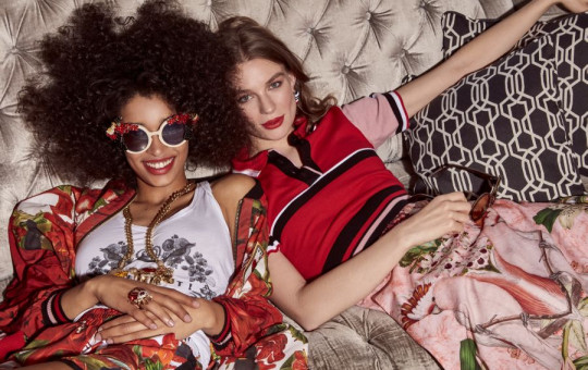 Two girls, where one is wearing sunglasses, are lying on a silver couch, wearing red, white and pink clothes, and smiling at the camera.