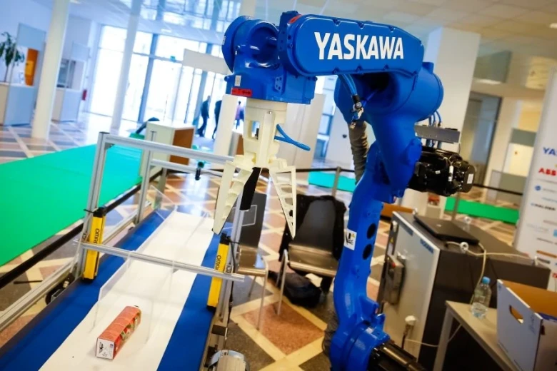 A blue Yaskawa industrial robot behind a conveyor belt that has a yoghurt in a red packaging on it. 