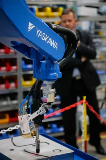 A blue Yaskawa robot and a man in the background, standing behind a red and white chain barrier.